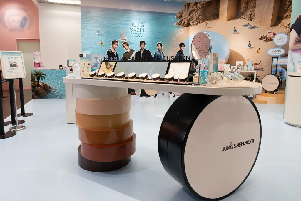 JUNG SAEM MOOLのクッションファンデーションを模した、特徴的なディスプレイカウンター。テスター、ミラー、サンゴの装飾を組み合わせた展示。背後にはビーチテーマのパネル。