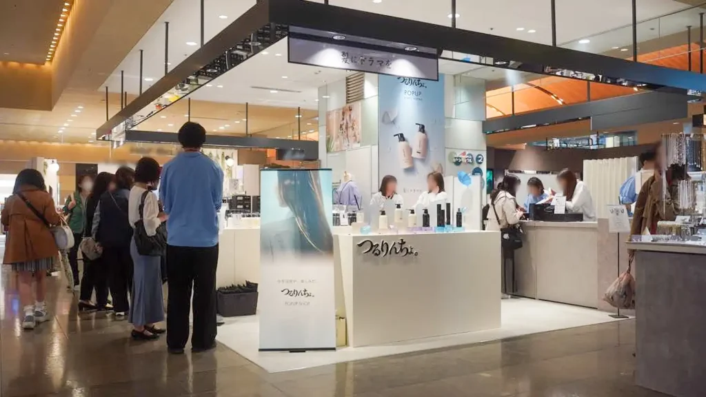 case_kaminidoramawo-03_02 | 株式会社ビーツ(BEEATS) Cosmetics kiosk in a shopping mall with staff behind a white counter and customers lined up early in the aisle.