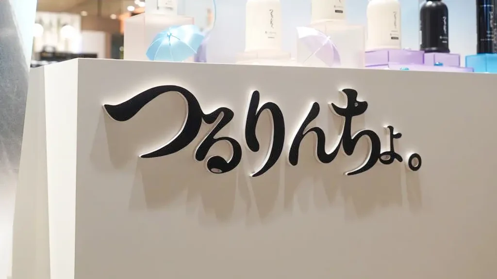 case_kaminidoramawo-03_03 | 株式会社ビーツ(BEEATS) Close-up of a white display counter with a black cursive logo mounted on it, with pastel beauty products visible on top behind the sign.