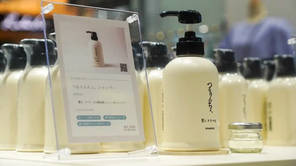 case_kaminidoramawo-03_04 | 株式会社ビーツ(BEEATS) Display of ivory shampoo bottles with black pumps lined up on a store shelf, with a small jar and a signboard in the foreground (Japanese text).