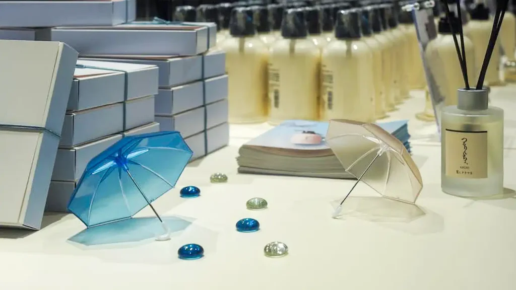 case_kaminidoramawo-03_07 | 株式会社ビーツ(BEEATS) Product display on a white surface with blue and clear miniature umbrellas, glass gems, stacked white boxes, a brochure, and a frosted diffuser bottle with sticks.